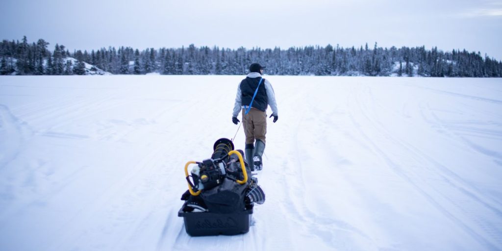 Image shows a man walking on a frozen lake while pulling a sled full of ice fishing gear. Used to promote ice fishing as one of the best Whiteshell Winter Activities