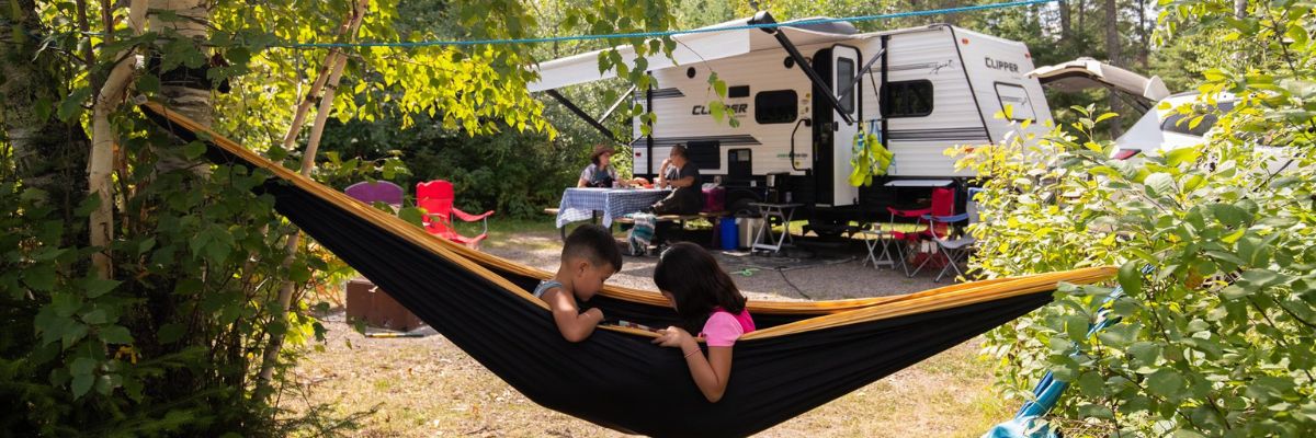 Two children in a hammock with a camping trailer and their parents in the background, showcasing the beauty and quality time families can have at Whiteshell campgrounds.
